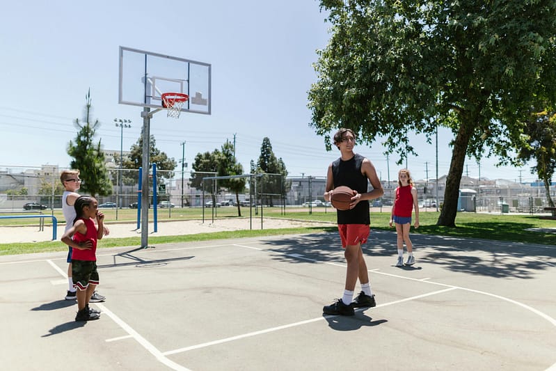 Children playing basketball on an outdoor court on a sunny day, with a hoop and tall tree in the background.